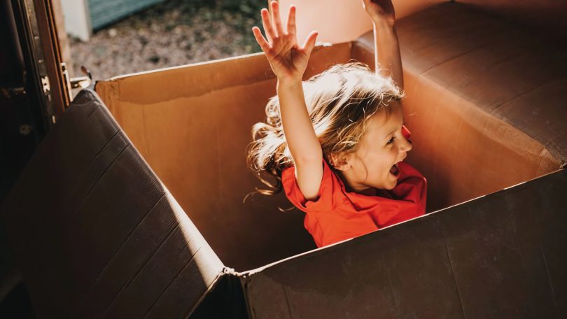 Niña feliz saltando y gritando de alegría dentro de una caja de cartón grande con las manos en alto