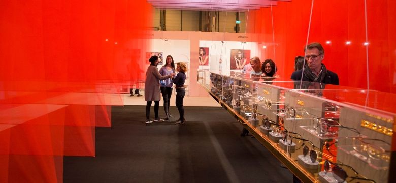 expositor de gafas de diseño en una vitrina de cristal durante la feria ExpoÓptica 2026 en IFEMA Madrid, con visitantes profesionales y una decoración de paneles rojos